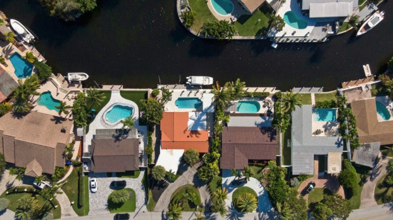 Aerial drone view of a luxury residential waterfront home in South Florida with a swimming pool, circular driveway, and lush landscaping.