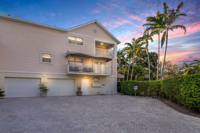 Beachfront vacation home with palm trees, sunset sky, and spacious driveway.