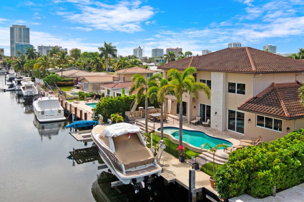 Aerial view of waterfront homes, boats, and lush greenery in a sunny cityscape.