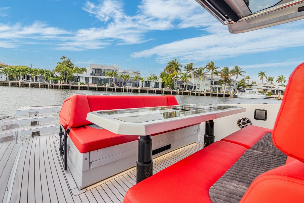 Outdoor yacht dining area with red cushioned seats and a table, overlooking a marina with palm trees.