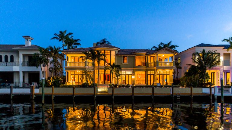 Luxury waterfront villa with palm trees, illuminated windows, and calm water reflections at dusk.
