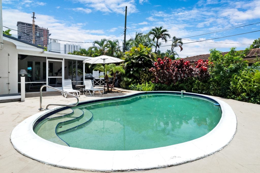 Poolside area with lounge chairs, umbrellas, and lush greenery in a sunny backyard.