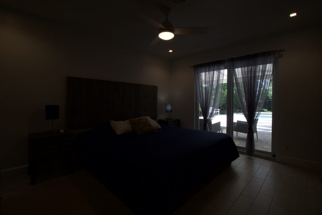 Dark bedroom with large window and curtains, minimal lighting, and ceiling fan.