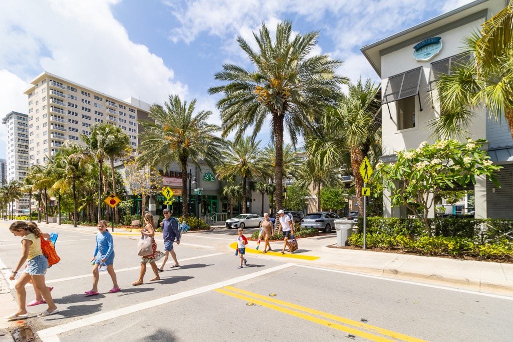 People crossing street in Pompano Beach with palm trees and shops.